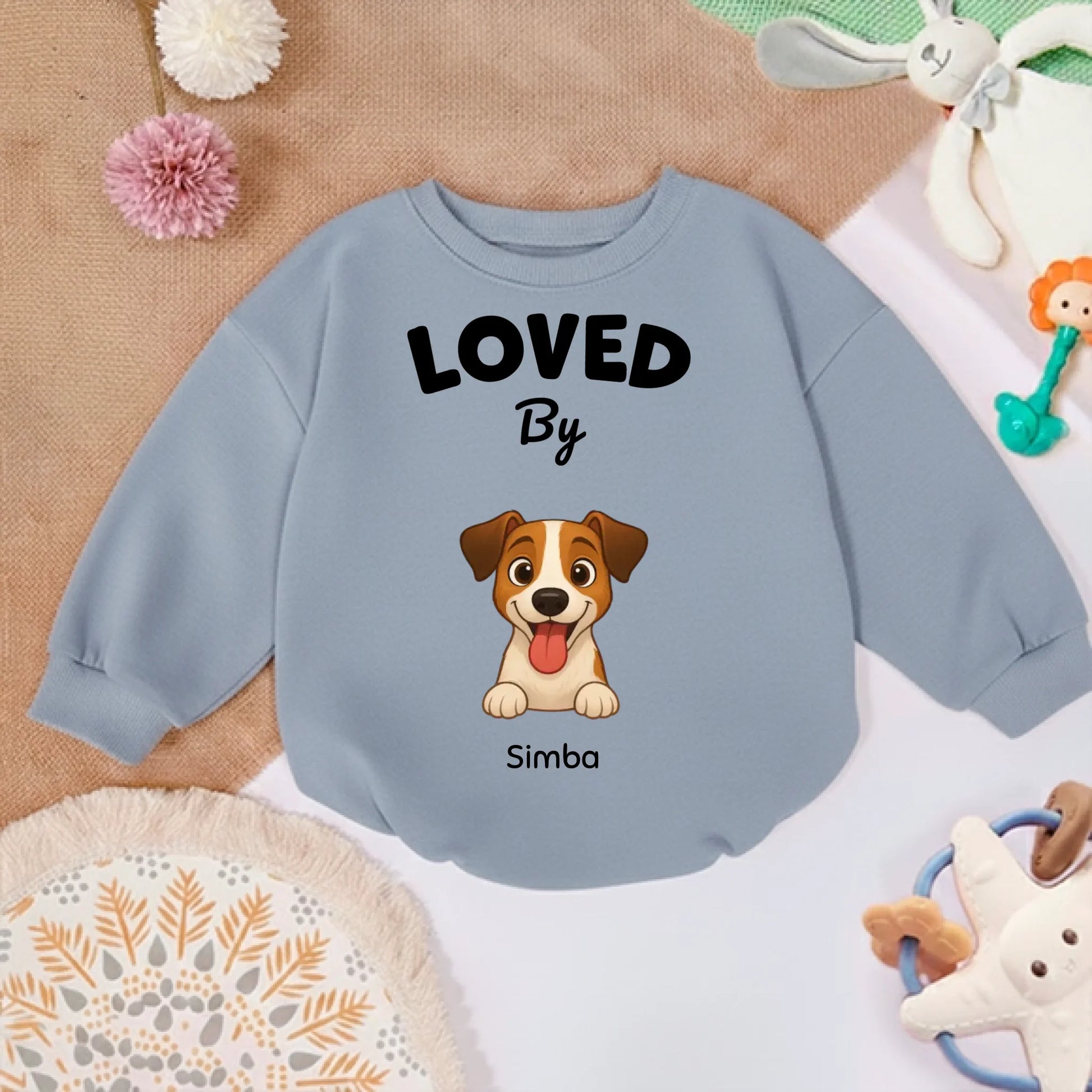 Pull bébé "LOVED By" posé sur un tapis doux avec des jouets pour enfant, ambiance chaleureuse.
