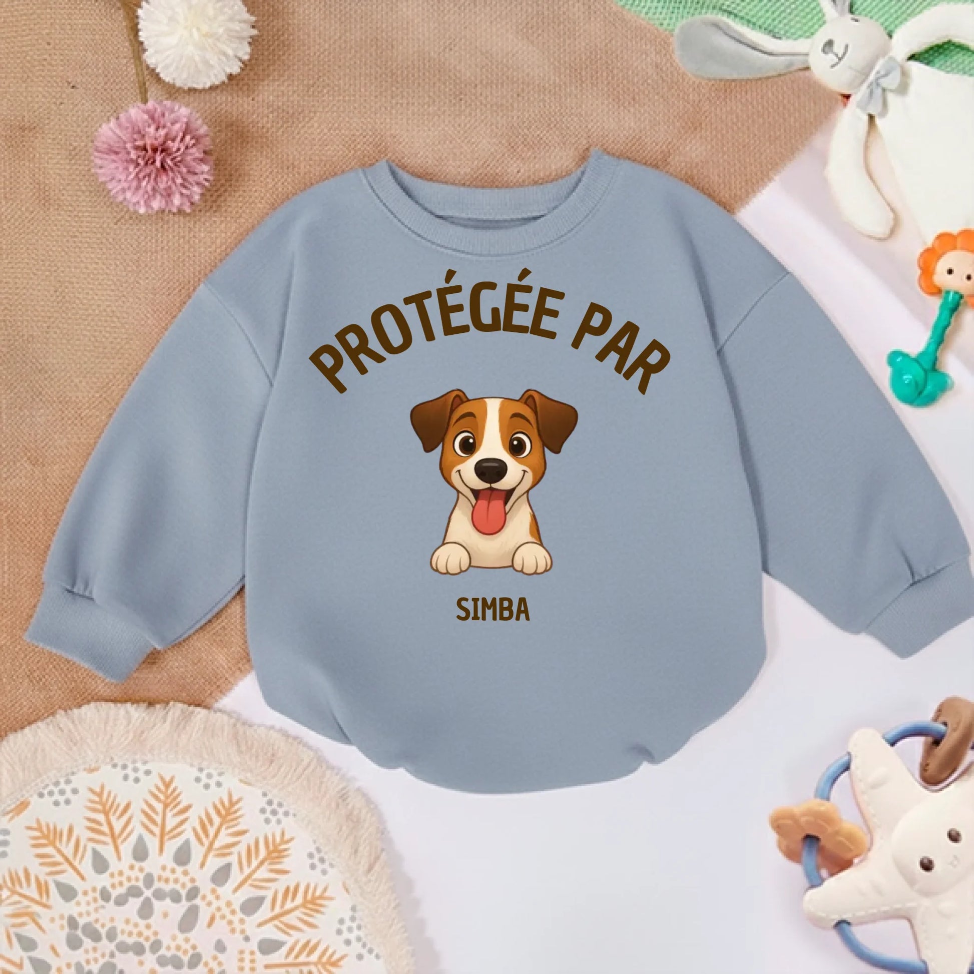 Pull bébé personnalisé posé sur un tapis doux avec des jouets pour bébé, suggérant un environnement de jeu.