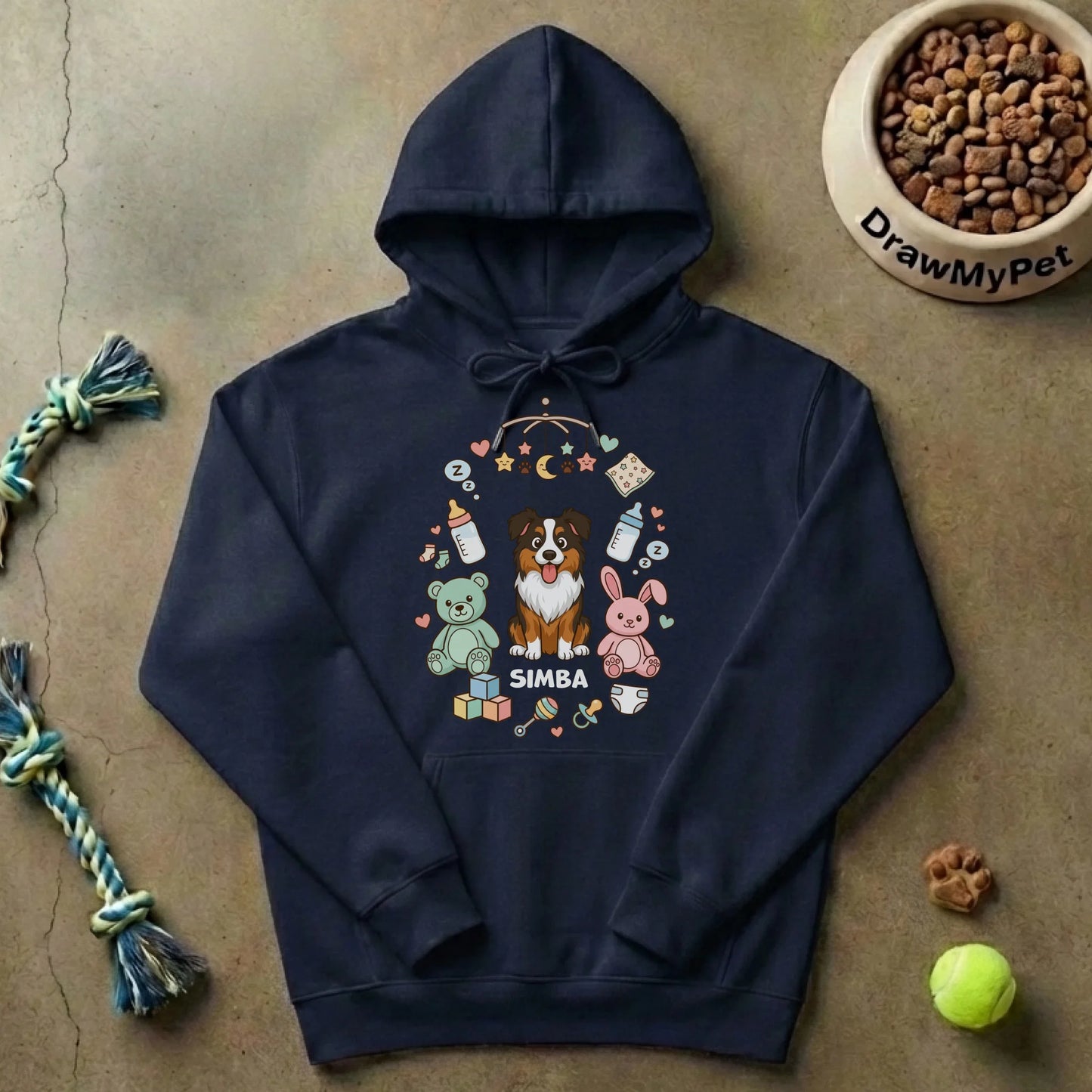 Pull à capuche personnalisé posé à plat sur un sol en béton, avec des jouets pour chien et une gamelle de croquettes, suggérant un environnement familial et animalier.