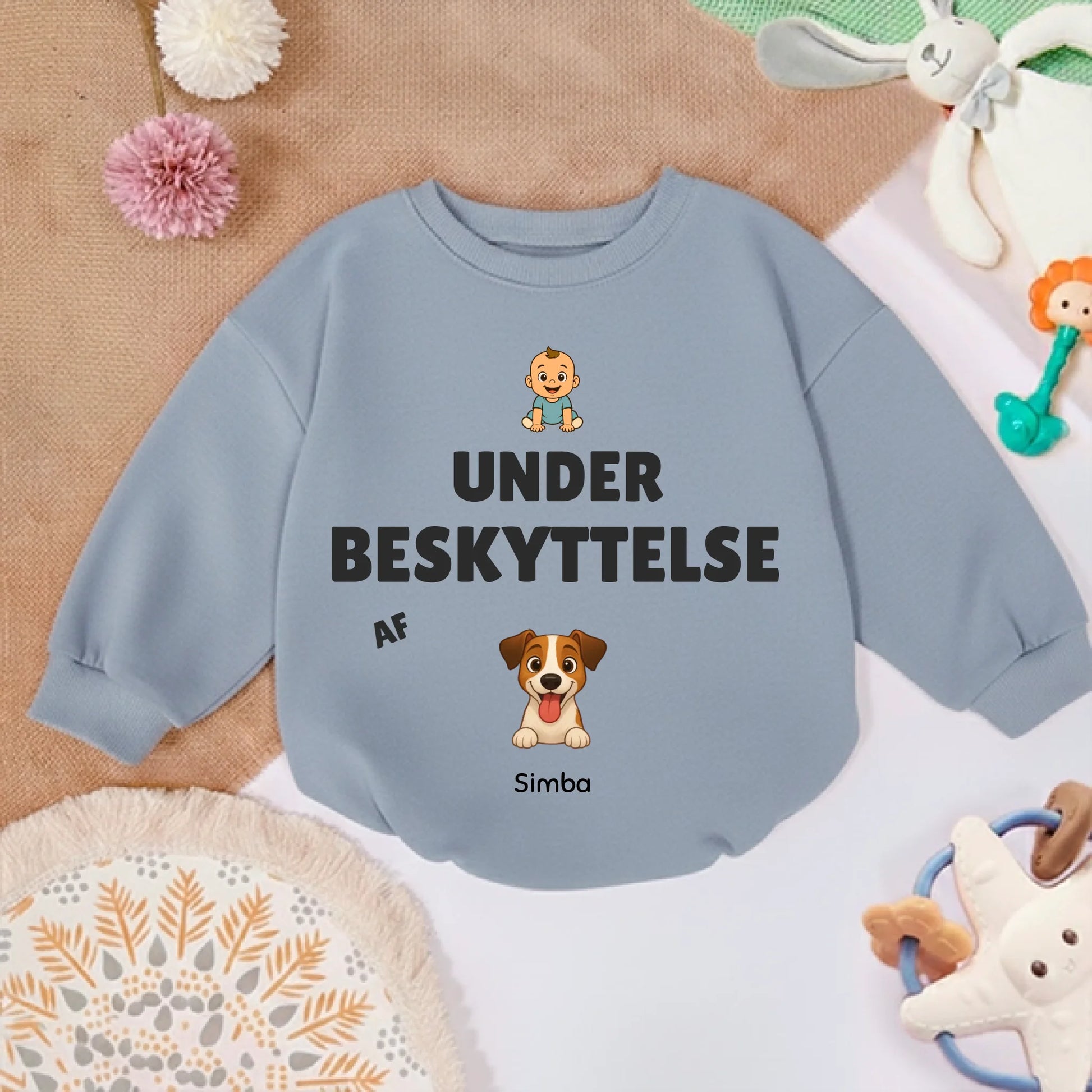Babytrøje 'Under beskyttelse af' placeret på et tæppe med legetøj, klar til brug.