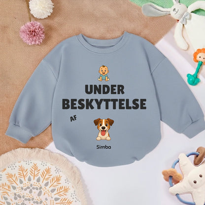 Babytrøje 'Under beskyttelse af' placeret på et tæppe med legetøj, klar til brug.