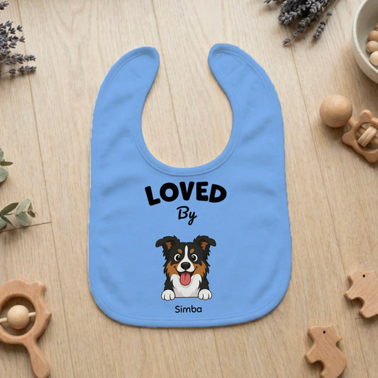 Personalized baby bib in light blue with 'LOVED By Simba' text and a happy cartoon dog portrait on a wooden background with baby toys.